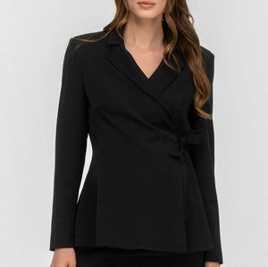 A woman modeling a black wrap-style blazer with a V-neck, notched lapels, long sleeves, and a self-tie waist, paired with black pants against a light gray studio background.
