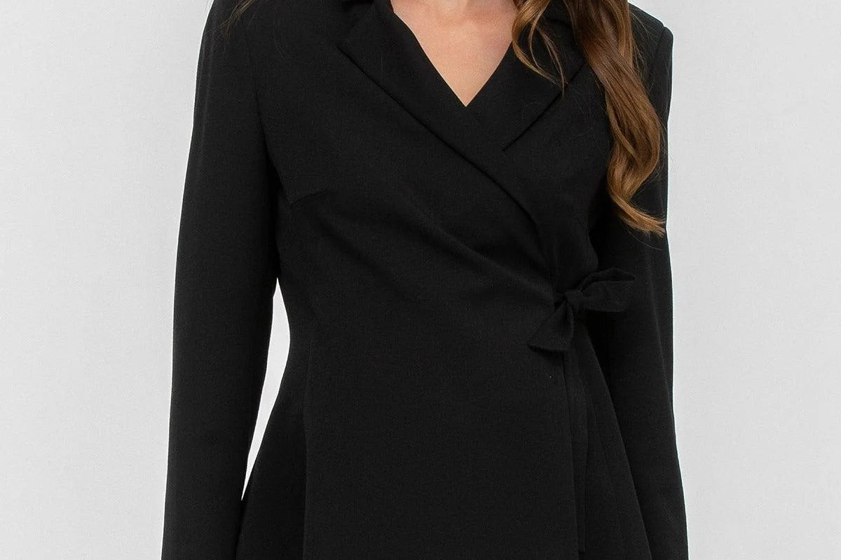 A woman modeling a black wrap-style blazer with a V-neck, notched lapels, long sleeves, and a self-tie waist, paired with black pants against a light gray studio background.