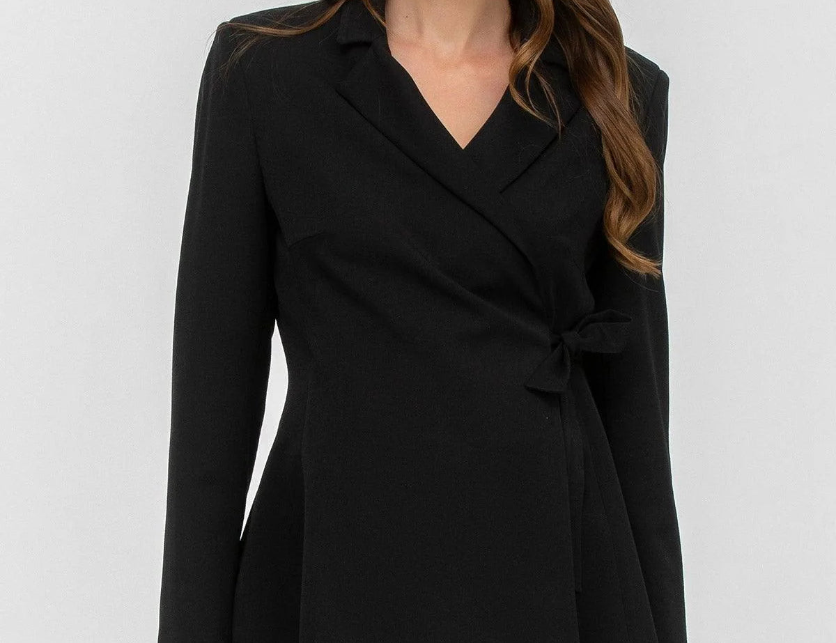 A woman modeling a black wrap-style blazer with a V-neck, notched lapels, long sleeves, and a self-tie waist, paired with black pants against a light gray studio background.