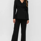 A woman modeling a black tailored two-piece suit consisting of a wrap-style blazer with peak lapels and matching wide-leg trousers, photographed against a clean light gray studio background.