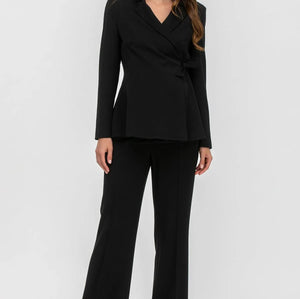 A woman modeling a black tailored two-piece suit consisting of a wrap-style blazer with peak lapels and matching wide-leg trousers, photographed against a clean light gray studio background.