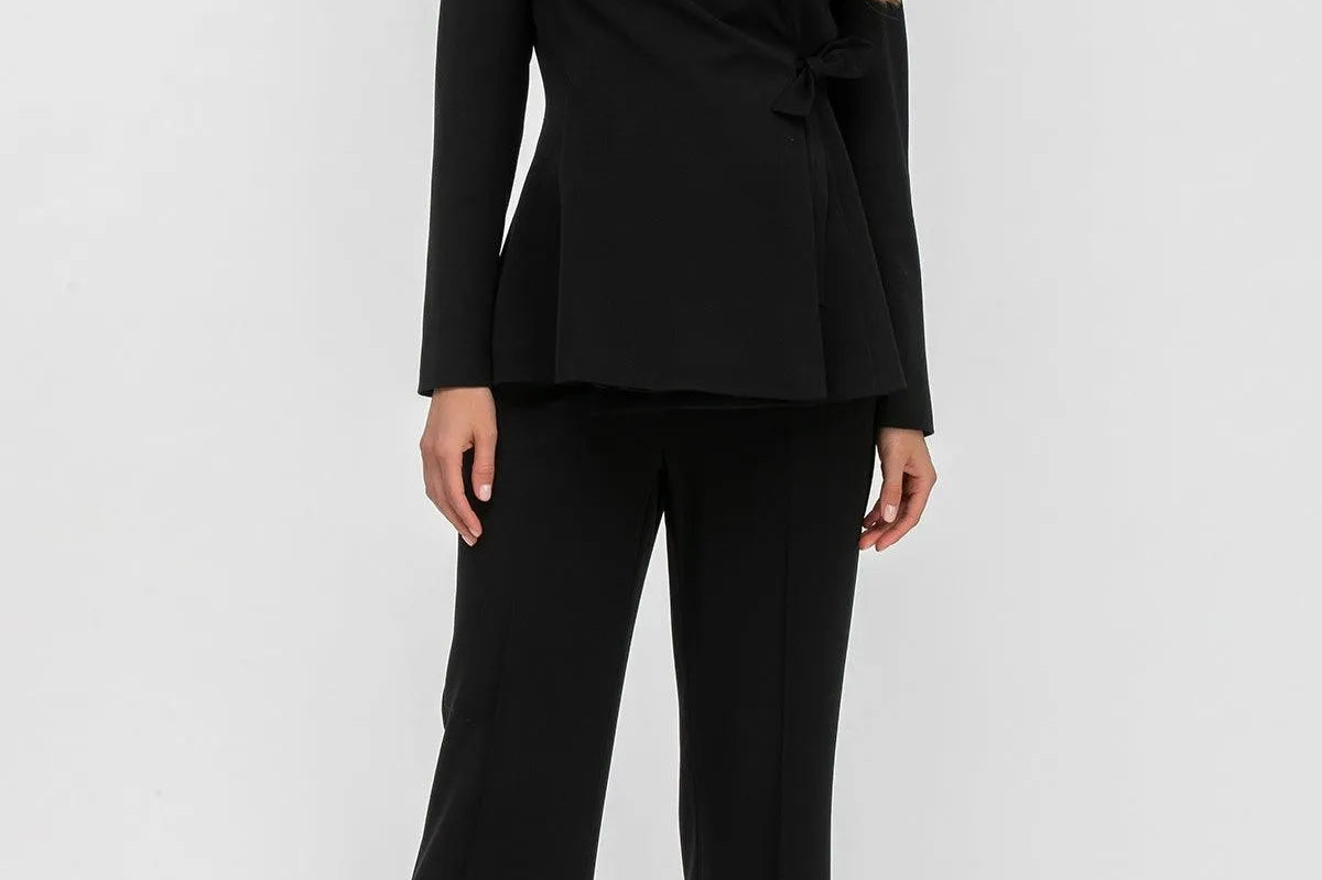 A woman modeling a black tailored two-piece suit consisting of a wrap-style blazer with peak lapels and matching wide-leg trousers, photographed against a clean light gray studio background.