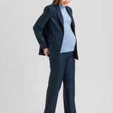 A pregnant woman modeling a navy blue maternity pantsuit, consisting of a single-breasted blazer and matching wide-leg trousers, worn over a light blue mock-neck top and brown pointed-toe heels, against a light gray studio background.