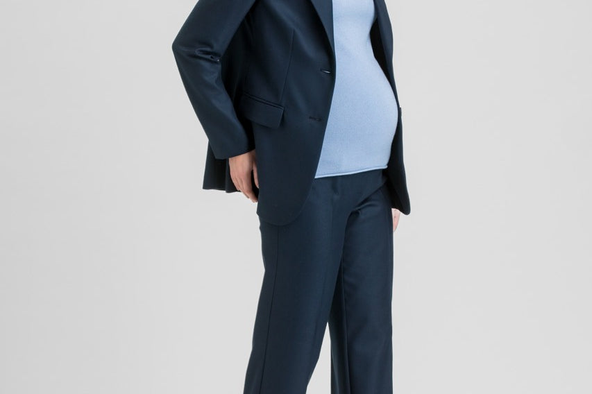 A pregnant woman modeling a navy blue maternity pantsuit, consisting of a single-breasted blazer and matching wide-leg trousers, worn over a light blue mock-neck top and brown pointed-toe heels, against a light gray studio background.