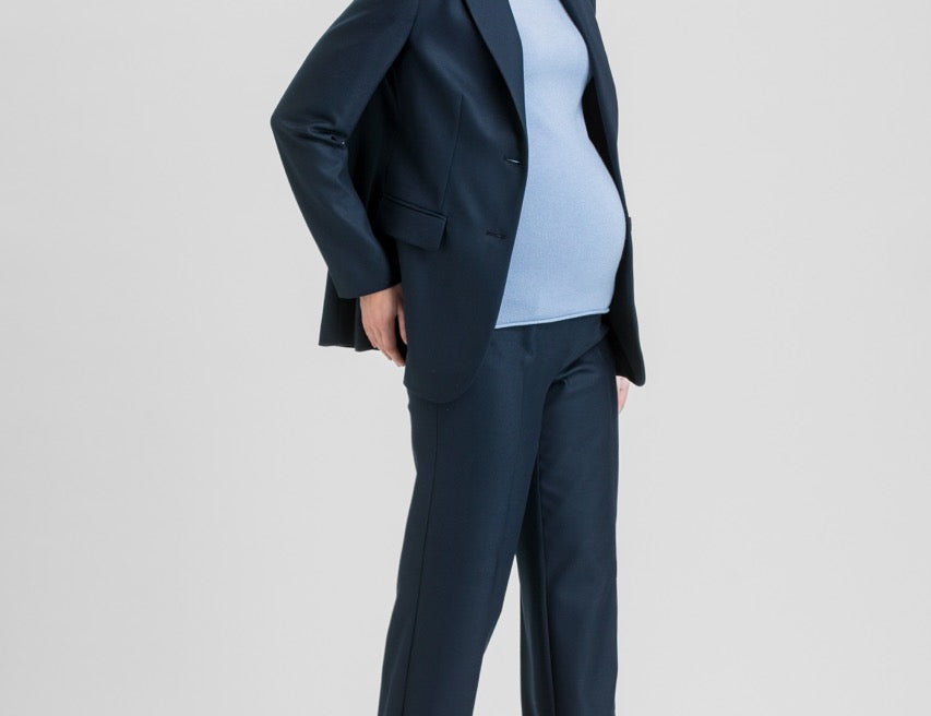 A pregnant woman modeling a navy blue maternity pantsuit, consisting of a single-breasted blazer and matching wide-leg trousers, worn over a light blue mock-neck top and brown pointed-toe heels, against a light gray studio background.