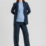 A woman modeling a navy blue tailored pantsuit, featuring a single-breasted blazer and matching wide-leg trousers, styled over a light blue mock-neck top and beige pointed-toe heels, against a solid light gray studio background.