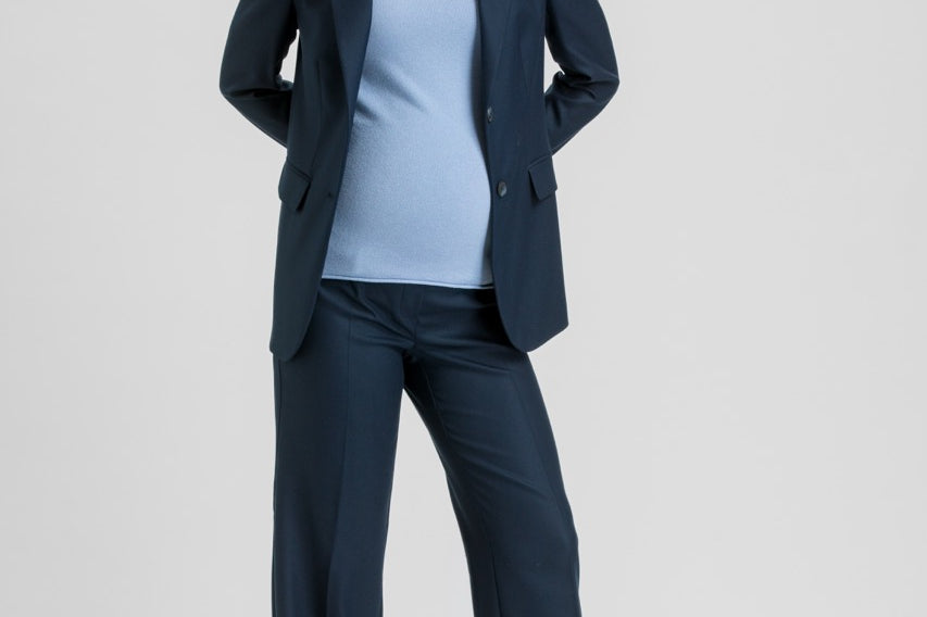 A woman modeling a navy blue tailored pantsuit, featuring a single-breasted blazer and matching wide-leg trousers, styled over a light blue mock-neck top and beige pointed-toe heels, against a solid light gray studio background.