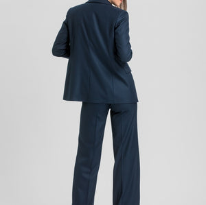 A woman modeling a navy blue two-piece suit from the back, featuring a tailored blazer and matching wide-leg trousers, set against a plain light gray studio background.