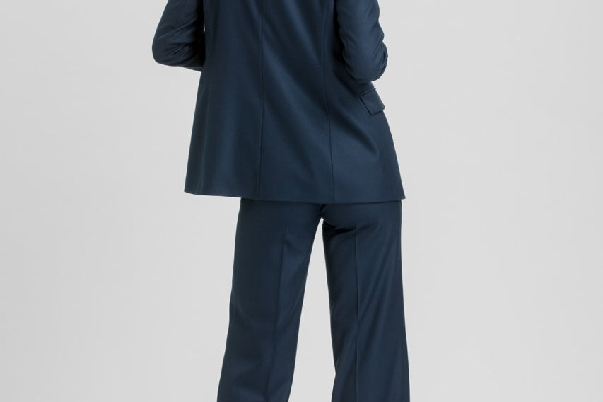 A woman modeling a navy blue two-piece suit from the back, featuring a tailored blazer and matching wide-leg trousers, set against a plain light gray studio background.