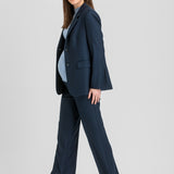 A pregnant model wearing a navy blue tailored pantsuit, featuring a single-breasted blazer and matching wide-leg trousers, styled with a light blue turtleneck and taupe pointed-toe heels against a light gray studio background.