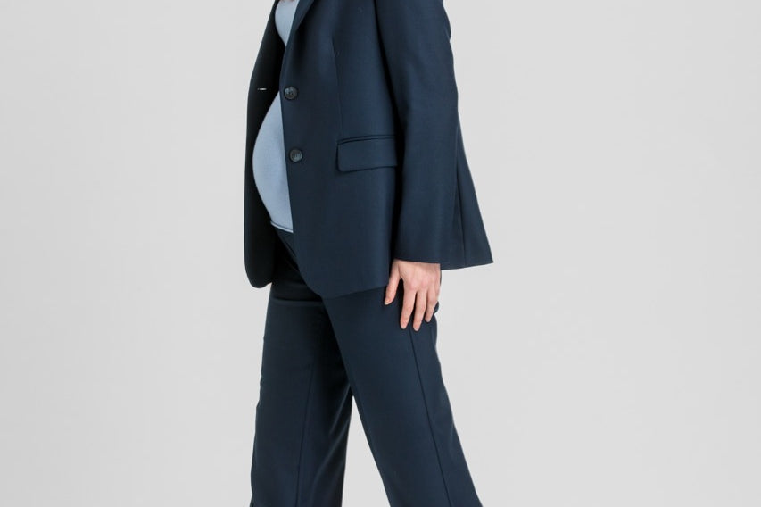 A pregnant model wearing a navy blue tailored pantsuit, featuring a single-breasted blazer and matching wide-leg trousers, styled with a light blue turtleneck and taupe pointed-toe heels against a light gray studio background.
