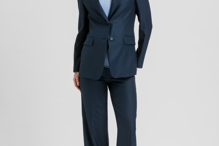 A woman modeling a navy blue two-piece suit with a light blue top and brown heels, standing against a light gray studio background.