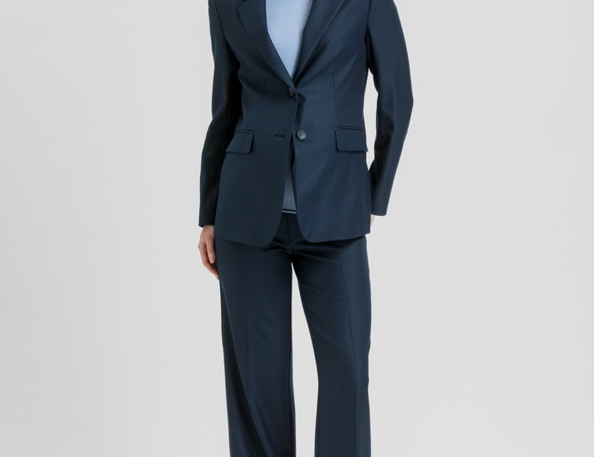A woman modeling a navy blue two-piece suit with a light blue top and brown heels, standing against a light gray studio background.