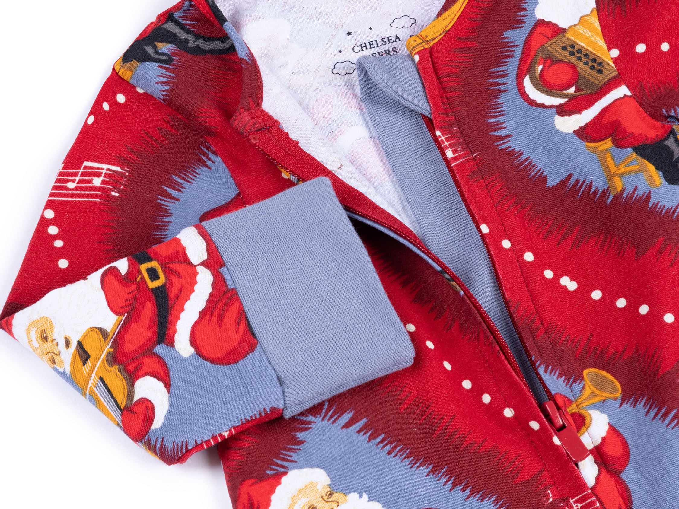 A close-up of a Christmas-themed zip-up pajama or long-sleeve top by Chelsea 1965, featuring a red and blue print of Santa Claus playing musical instruments, with a solid blue cuff.