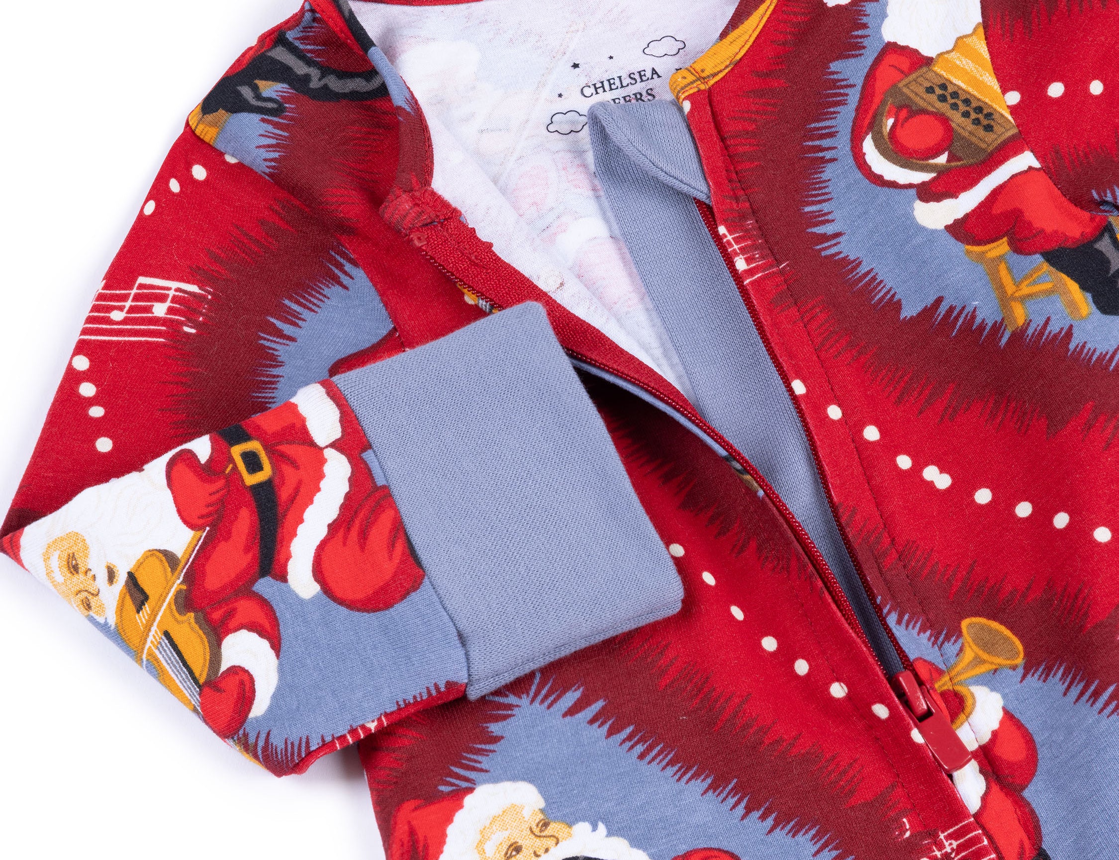 A close-up of a Christmas-themed zip-up pajama or long-sleeve top by Chelsea 1965, featuring a red and blue print of Santa Claus playing musical instruments, with a solid blue cuff.