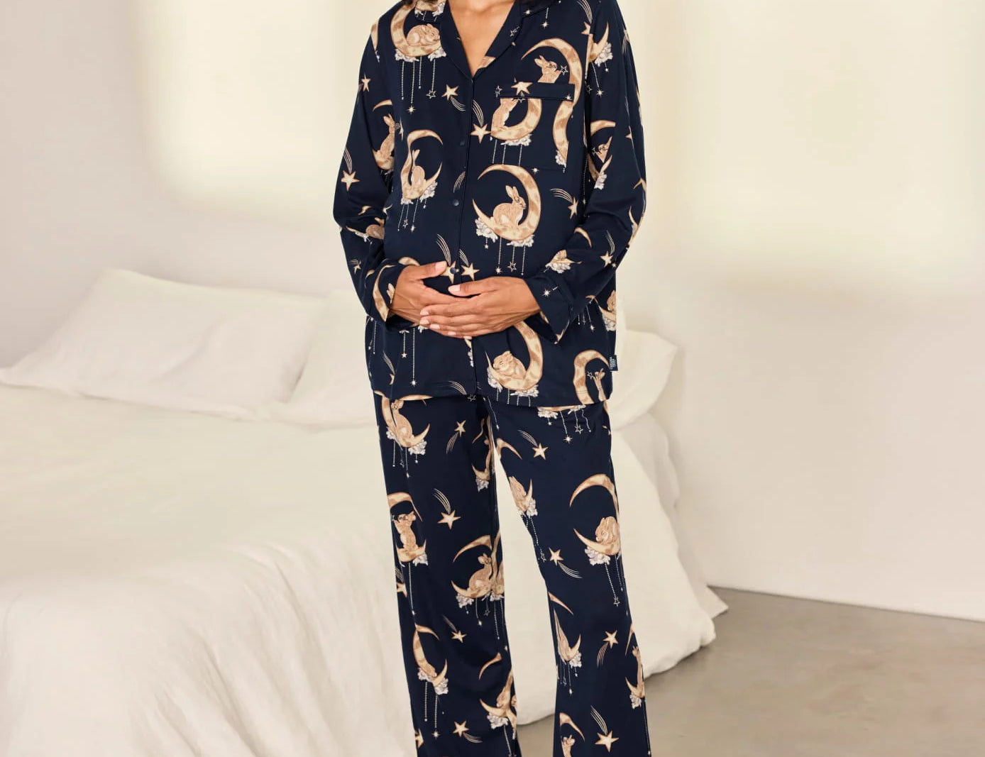 A woman stands barefoot in a bedroom wearing a navy blue pajama set with a beige cat-and-moon print. The set includes a long-sleeve button-front top and matching wide-leg pants.