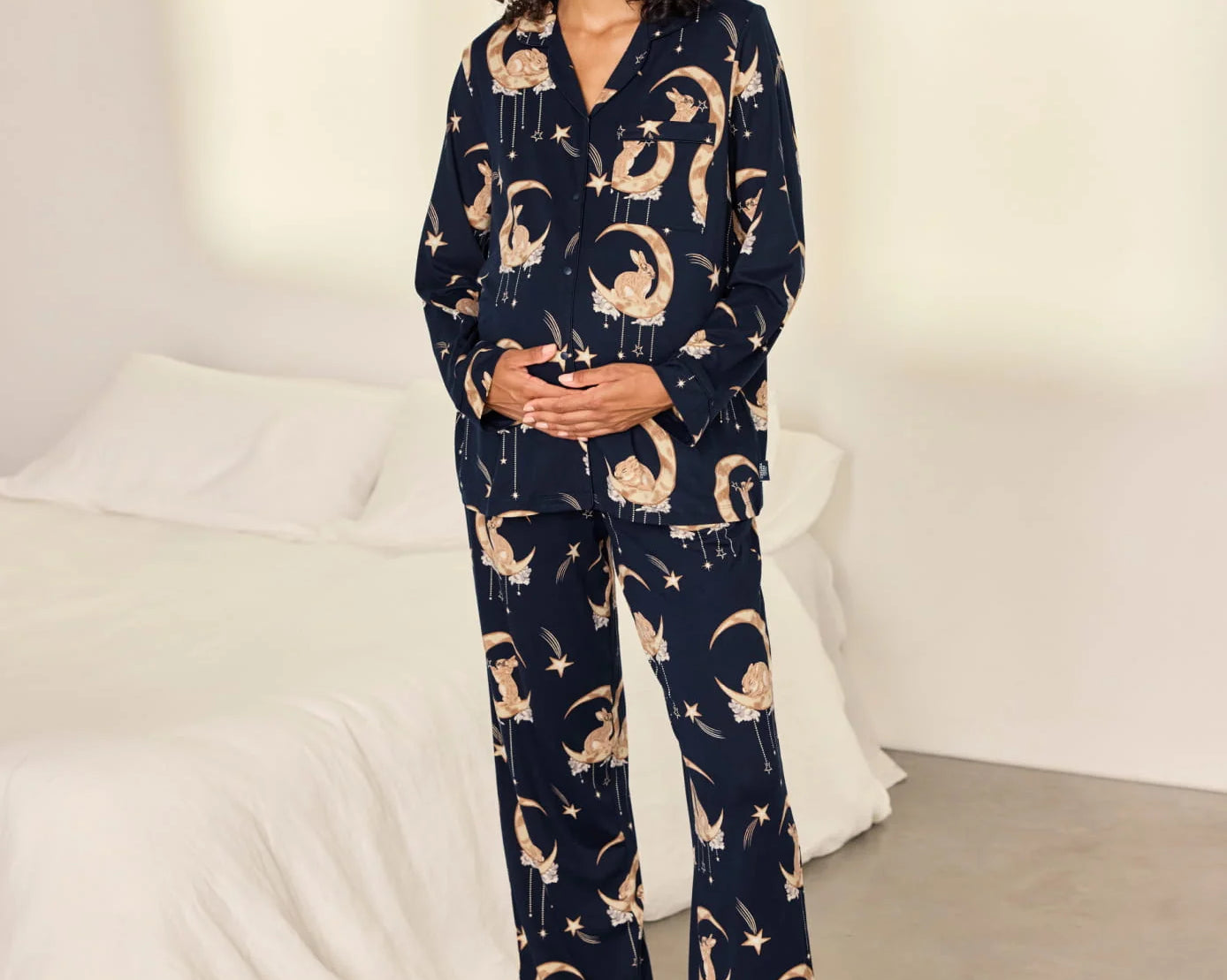 A woman stands barefoot in a bedroom wearing a navy blue pajama set with a beige cat-and-moon print. The set includes a long-sleeve button-front top and matching wide-leg pants.