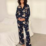 A woman stands barefoot in a bedroom wearing a navy blue pajama set with a beige cat-and-moon print. The set includes a long-sleeve button-front top and matching wide-leg pants.