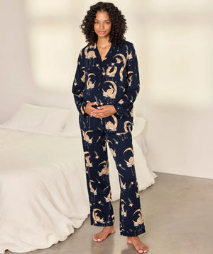 A woman stands barefoot in a bedroom wearing a navy blue pajama set with a beige cat-and-moon print. The set includes a long-sleeve button-front top and matching wide-leg pants.