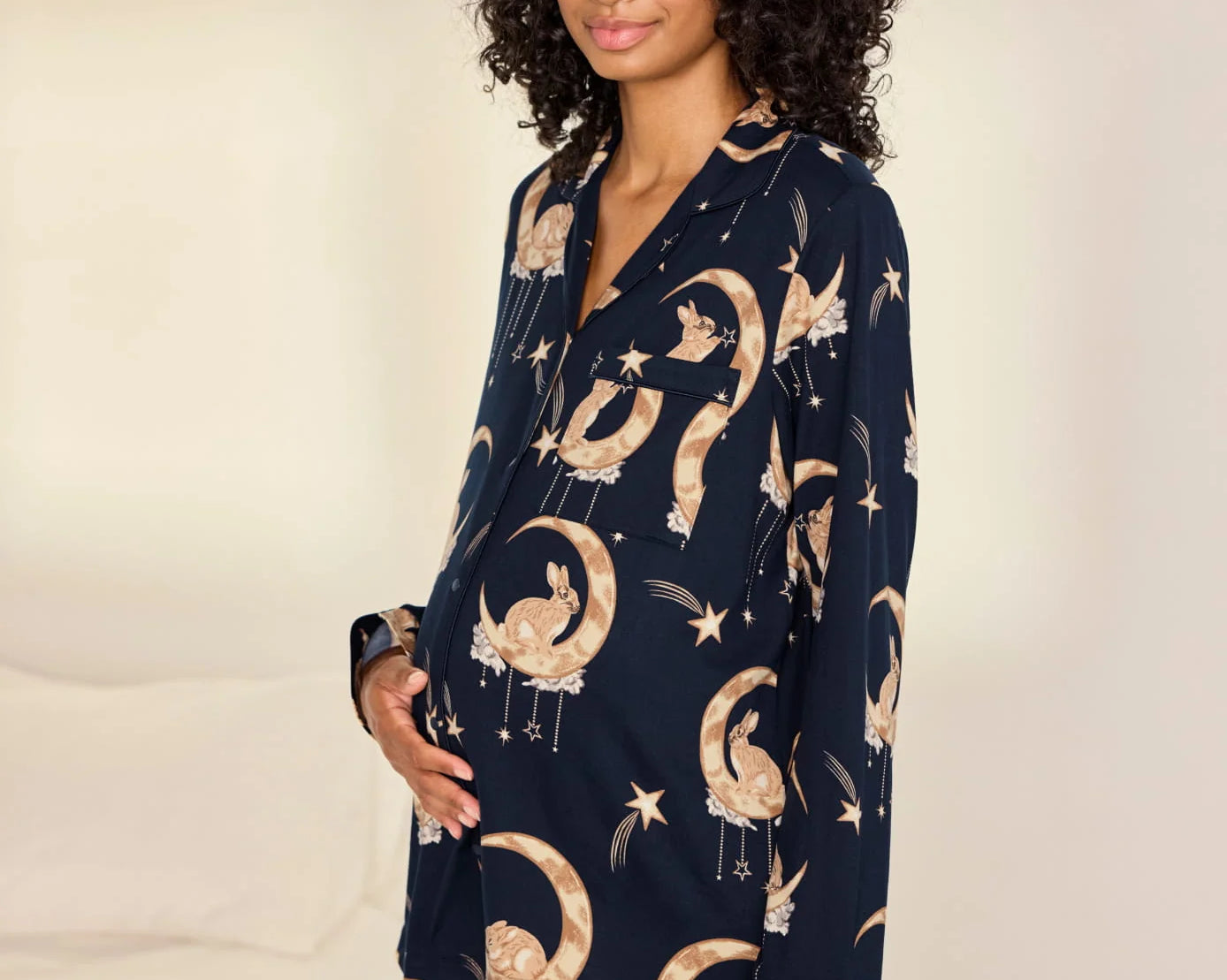 A woman modeling a navy blue pajama set with a celestial print of gold crescent moons, stars, and birds, set against a warm off-white background.