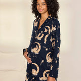 A woman modeling a navy blue pajama set with a celestial print of gold crescent moons, stars, and birds, set against a warm off-white background.
