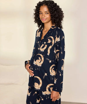 A woman modeling a navy blue pajama set with a celestial print of gold crescent moons, stars, and birds, set against a warm off-white background.