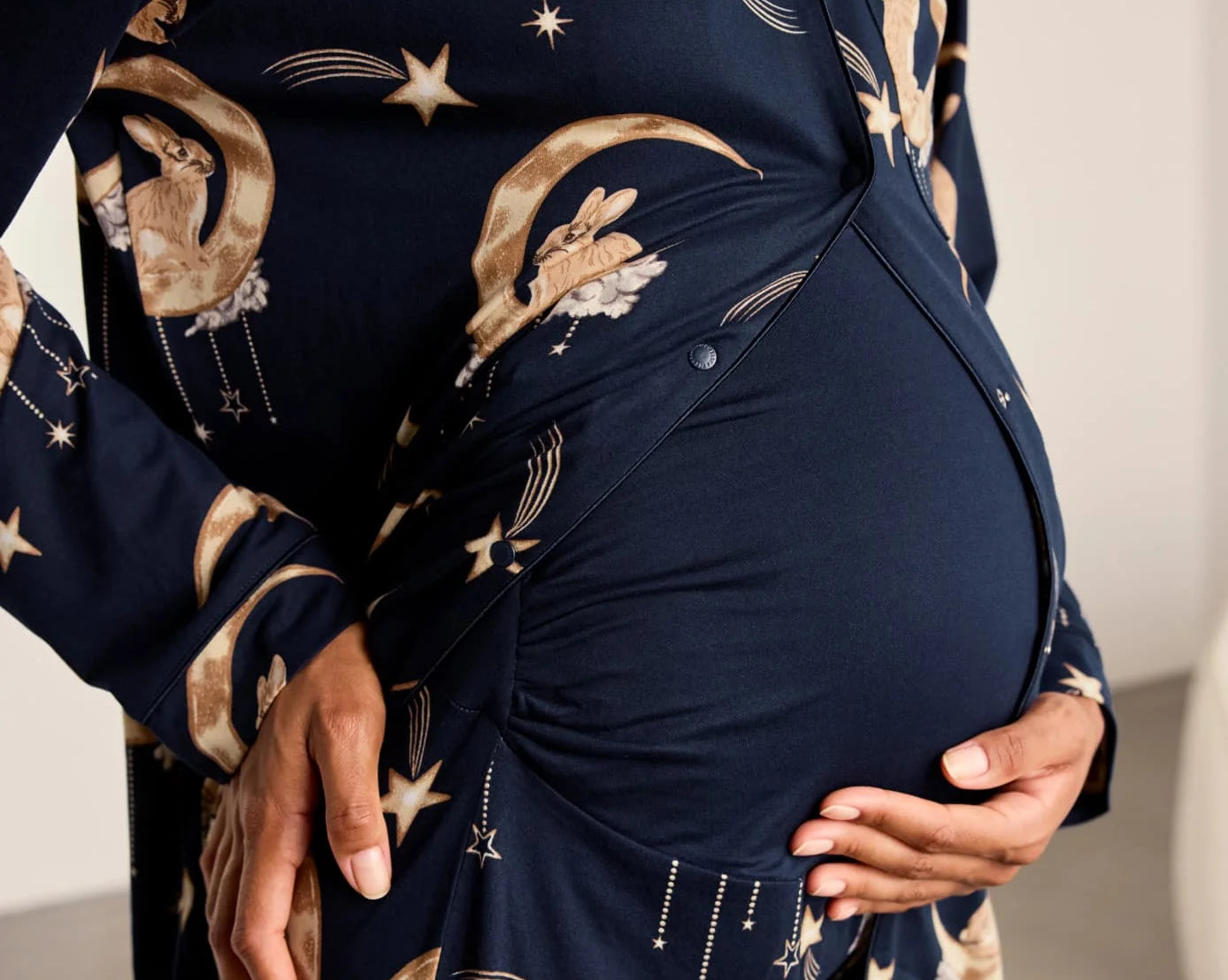 A close-up of a pregnant model wearing a navy blue long-sleeve maternity sleep shirt or robe with a celestial print of gold stars, moons, and rabbits, styled with a matching belly wrap.