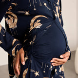 A close-up of a pregnant model wearing a navy blue long-sleeve maternity sleep shirt or robe with a celestial print of gold stars, moons, and rabbits, styled with a matching belly wrap.