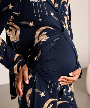 A close-up of a pregnant model wearing a navy blue long-sleeve maternity sleep shirt or robe with a celestial print of gold stars, moons, and rabbits, styled with a matching belly wrap.