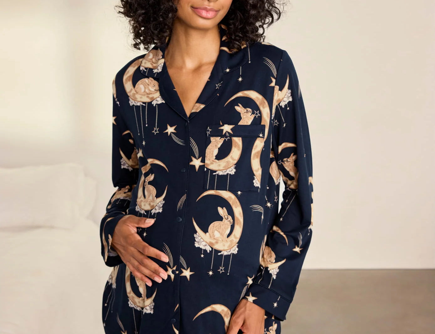 A woman modeling a navy blue pajama set with a celestial print of gold crescent moons, stars, and flowers, set against a neutral bedroom background.