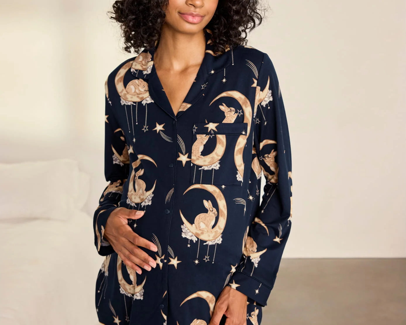 A woman modeling a navy blue pajama set with a celestial print of gold crescent moons, stars, and flowers, set against a neutral bedroom background.