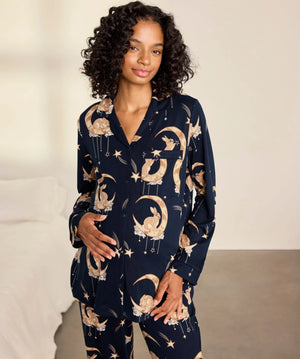 A woman modeling a navy blue pajama set with a celestial print of gold crescent moons, stars, and flowers, set against a neutral bedroom background.