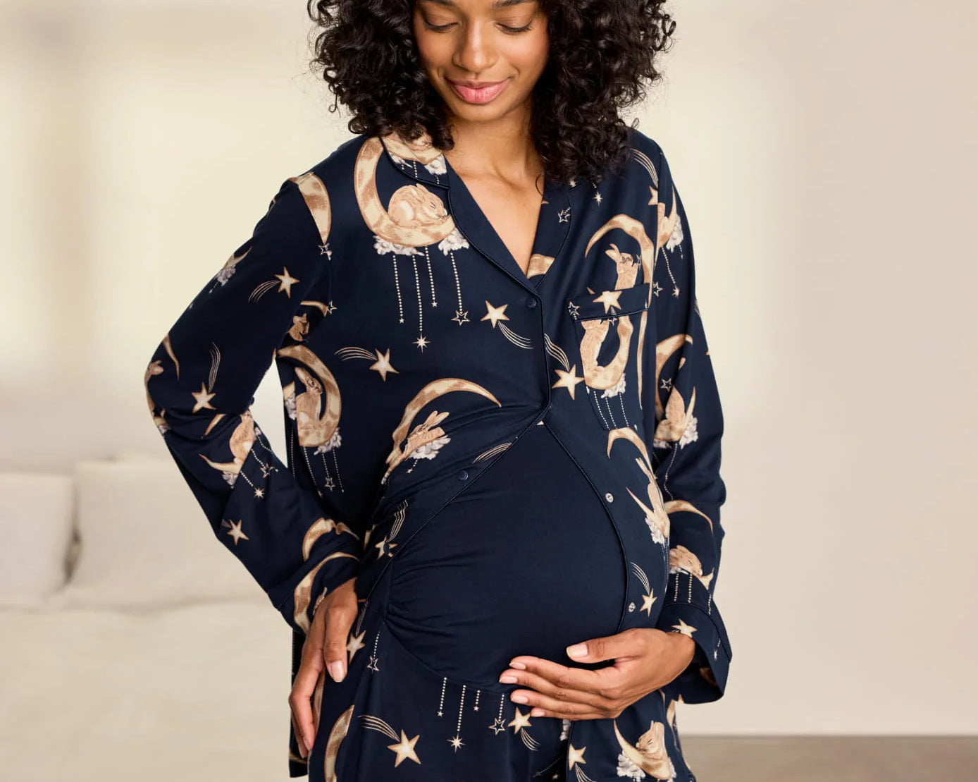 A pregnant woman wearing a navy blue maternity pajama set with a celestial print of gold crescent moons, stars, and rabbits, standing in a softly lit bedroom.