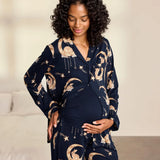 A pregnant woman wearing a navy blue maternity pajama set with a celestial print of gold crescent moons, stars, and rabbits, standing in a softly lit bedroom.