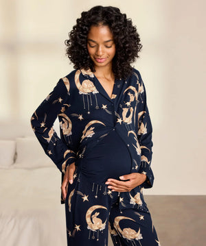 A pregnant woman wearing a navy blue maternity pajama set with a celestial print of gold crescent moons, stars, and rabbits, standing in a softly lit bedroom.