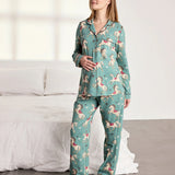 A woman modeling a two-piece pajama set with a teal green background and a whimsical pattern of pink, red, and white birds, standing in a brightly lit bedroom next to a bed with white linens.