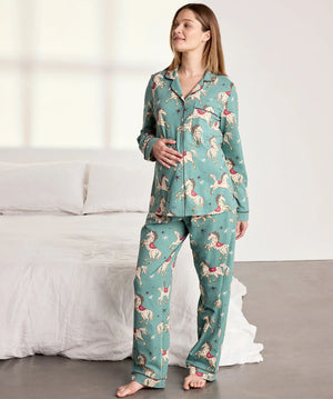 A woman modeling a two-piece pajama set with a teal green background and a whimsical pattern of pink, red, and white birds, standing in a brightly lit bedroom next to a bed with white linens.