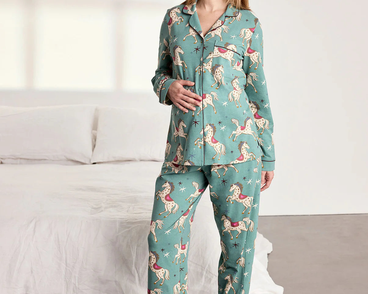 A woman modeling a two-piece pajama set with a teal green background and a whimsical pattern of pink, red, and white birds, standing in a brightly lit bedroom next to a bed with white linens.