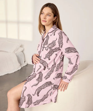 A woman wearing a pink long-sleeve pajama shirt dress with a black leopard print pattern, sitting on a white armchair in a bright bedroom setting.