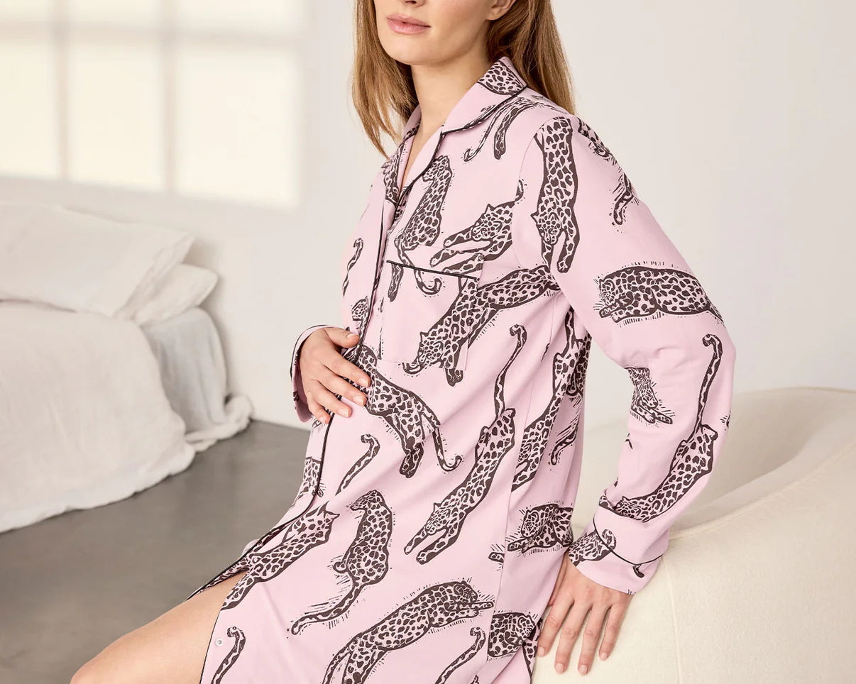 A woman wearing a pink long-sleeve pajama shirt dress with a black leopard print pattern, sitting on a white armchair in a bright bedroom setting.