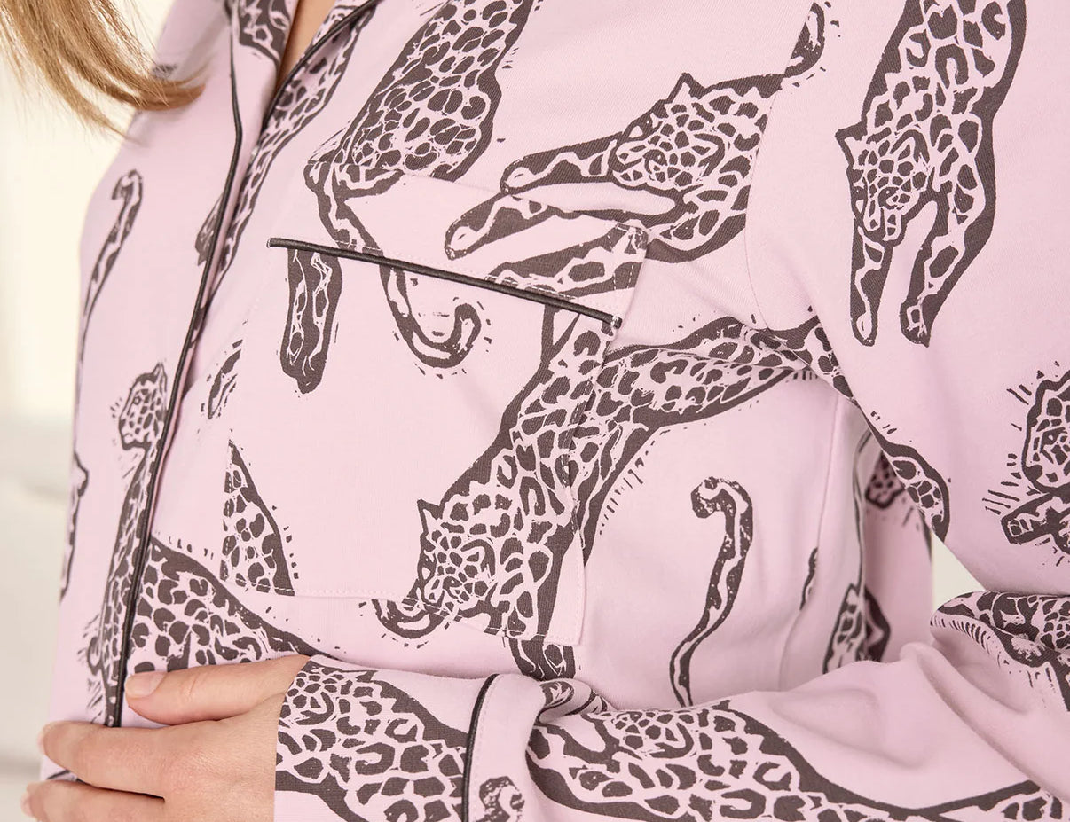 A close-up of a person wearing a light pink long-sleeve pajama shirt with a dark brown giraffe print and black piping along the collar, placket, and pocket.