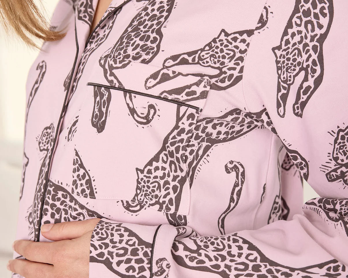 A close-up of a person wearing a light pink long-sleeve pajama shirt with a dark brown giraffe print and black piping along the collar, placket, and pocket.