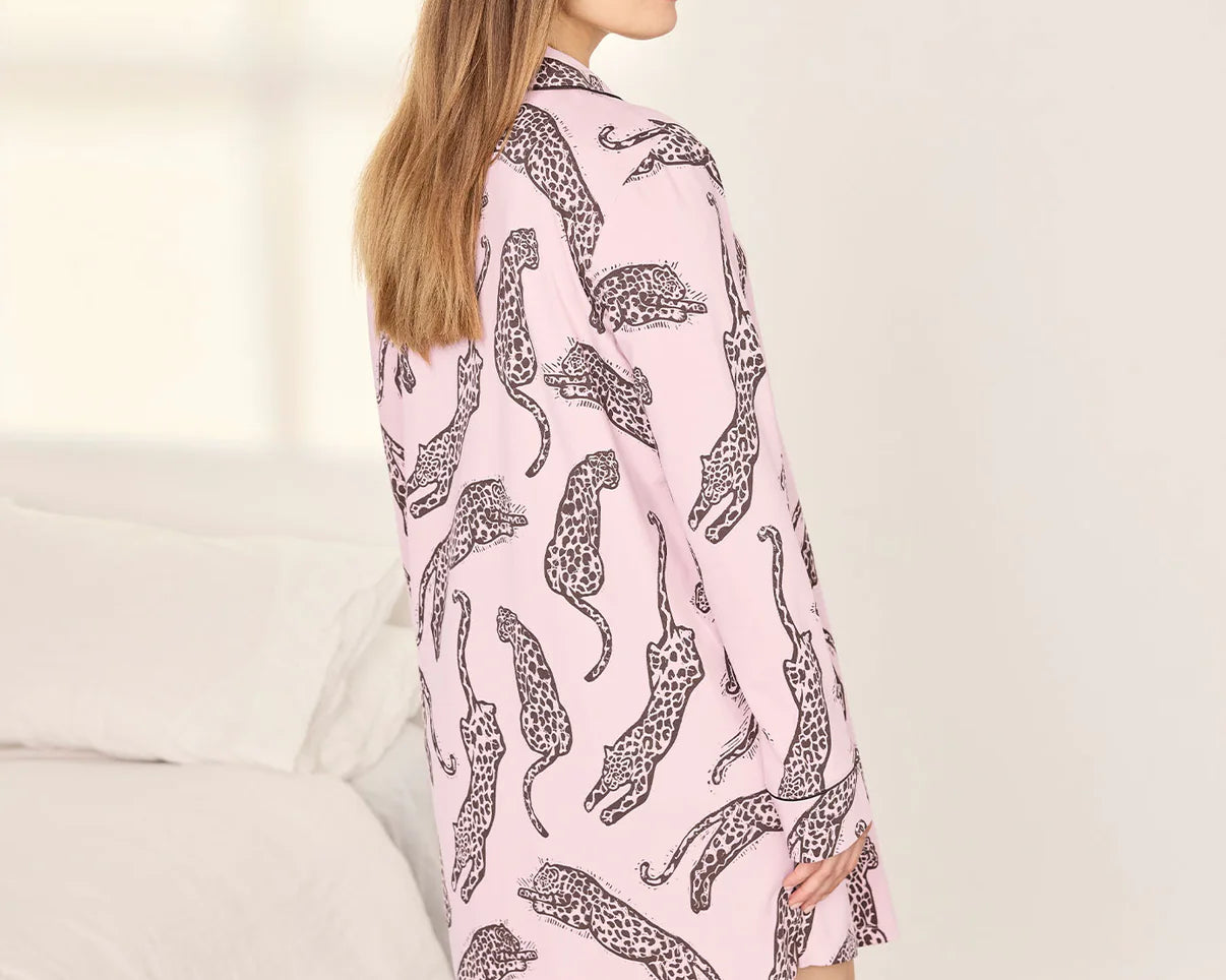 A woman wearing a pink long-sleeve shirt dress with a black leopard print pattern, standing in a brightly lit bedroom.