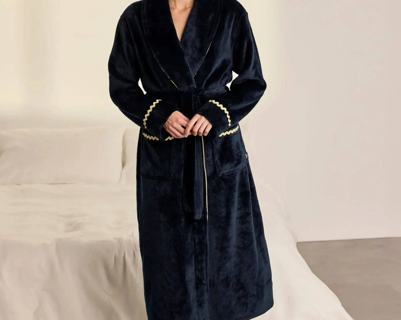 A woman wearing a plush navy blue robe with gold trim and a gold belt, standing barefoot in a bedroom setting with a bed and white sheets in the background.