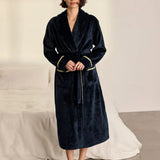 A woman wearing a plush navy blue robe with gold trim and a gold belt, standing barefoot in a bedroom setting with a bed and white sheets in the background.