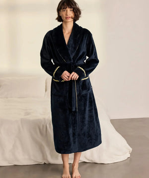 A woman wearing a plush navy blue robe with gold trim and a gold belt, standing barefoot in a bedroom setting with a bed and white sheets in the background.