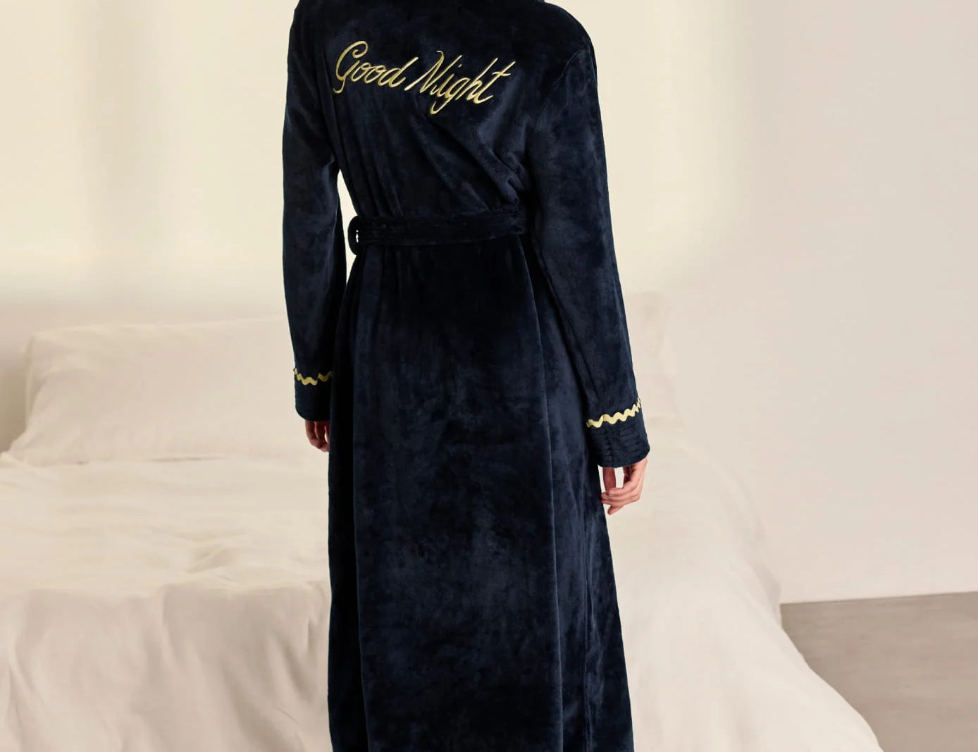 A woman stands with her back to the camera wearing a dark navy blue plush bathrobe with gold trim and 'Good Night' embroidered on the back, in a bedroom setting with a bed in the background.