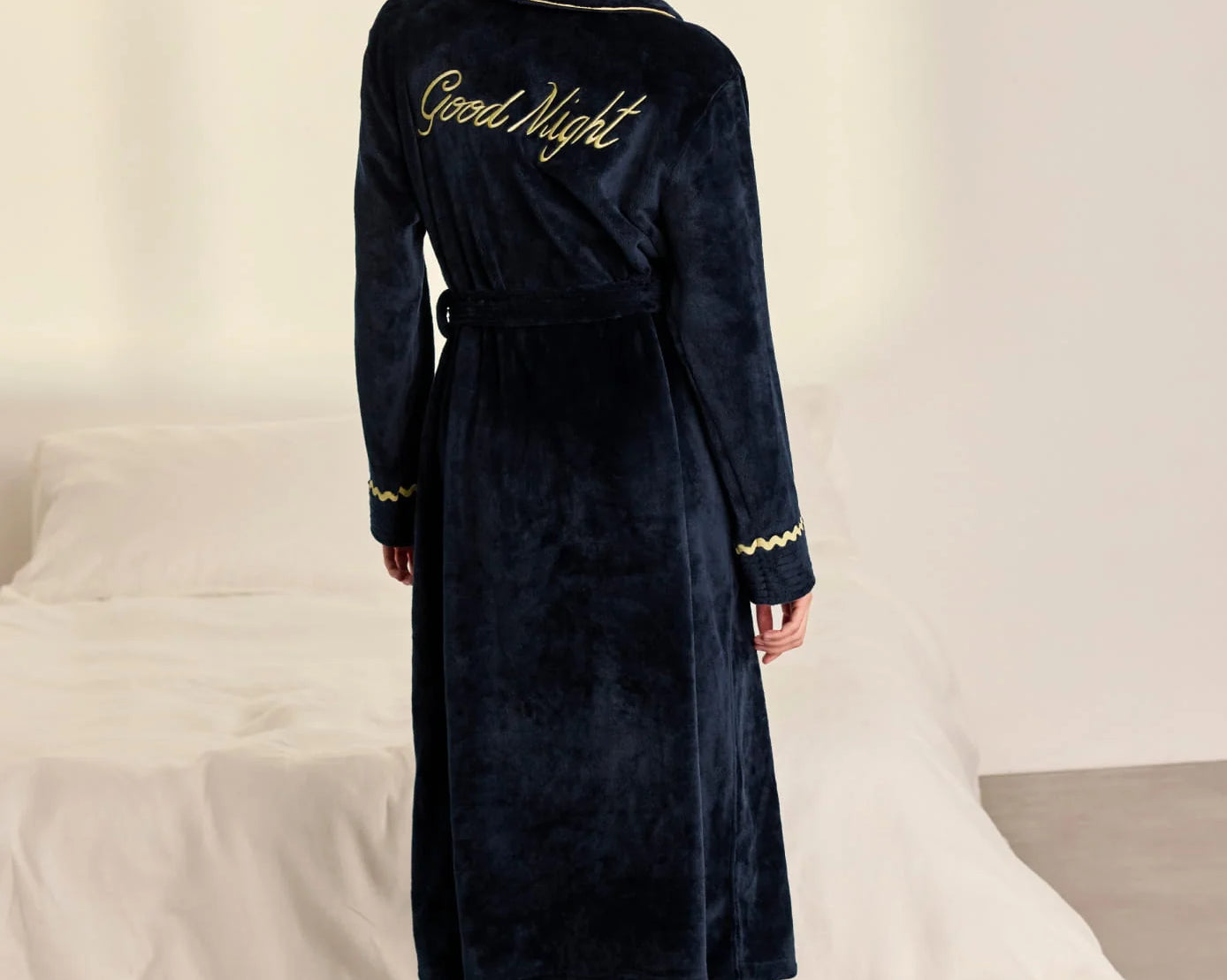 A woman stands with her back to the camera wearing a dark navy blue plush bathrobe with gold trim and 'Good Night' embroidered on the back, in a bedroom setting with a bed in the background.