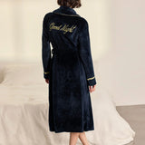 A woman stands with her back to the camera wearing a dark navy blue plush bathrobe with gold trim and 'Good Night' embroidered on the back, in a bedroom setting with a bed in the background.
