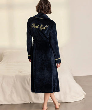 A woman stands with her back to the camera wearing a dark navy blue plush bathrobe with gold trim and 'Good Night' embroidered on the back, in a bedroom setting with a bed in the background.